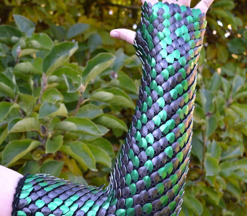 A photo of an arm wearing a grean and black scaly sleeve made of knitted scalemaille by Crystal's Idyll.