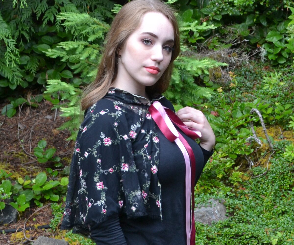 A black capelet wiht floral patterns and long pink and maroon ribbon, hood down, modelled by a young woman.